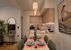 A modern dining area with a wooden table set for four, a kitchen in the background, and a large mirror on the wall. - LIV - Chic 2BR Signature Flat | Near Metro (Athens)