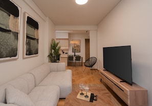 A modern living room with a sectional sofa, a flat-screen TV on a wooden stand, and a dining area in the background. - LIV - Chic 2BR Signature Flat | Near Metro (Athens)