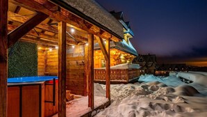 Eine Holzhütte mit überdachter Außensitzgruppe und verschneierter Landschaft bei Nacht.