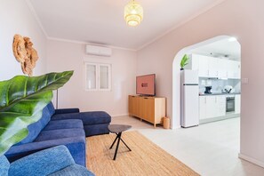 Ein modernes Wohnzimmer mit einem blauen Sofa, einer großen Zimmerpflanze und einem kleinen Beistelltisch. Ein Holzkasten und ein Fernseher auf einem Schrank stehen dort. Das Zimmer geht in eine Küche mit weißen Schränken und einem Kühlschrank über.
