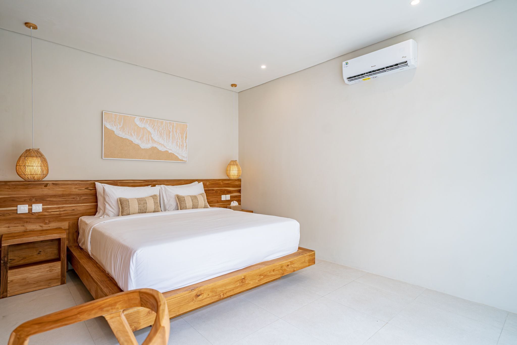A bedroom with a wooden bed frame, white bedding, a wall-mounted air conditioner, and a beach-themed artwork.