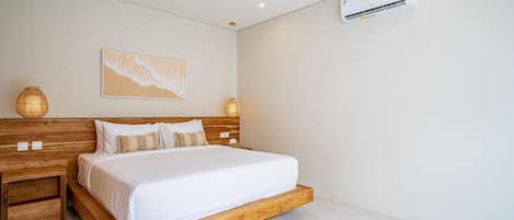 A bedroom with a wooden bed frame, white bedding, a wall-mounted air conditioner, and a beach-themed artwork.