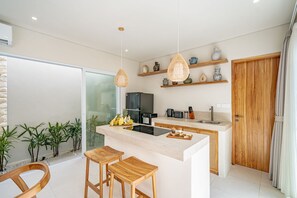 A modern kitchen with a central island, wooden stools, and a glass door leading to a balcony. - Rocha Villa Uluwatu (Uluwatu)