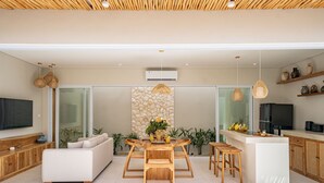 A modern kitchen with a central island, a dining area with a wooden table and chairs, and a living area with a sofa and television. - Rocha Villa Uluwatu (Uluwatu)