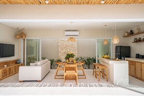 A modern kitchen with a central island, a dining area with a wooden table and chairs, and a living area with a sofa and television. - Rocha Villa Uluwatu (Uluwatu)