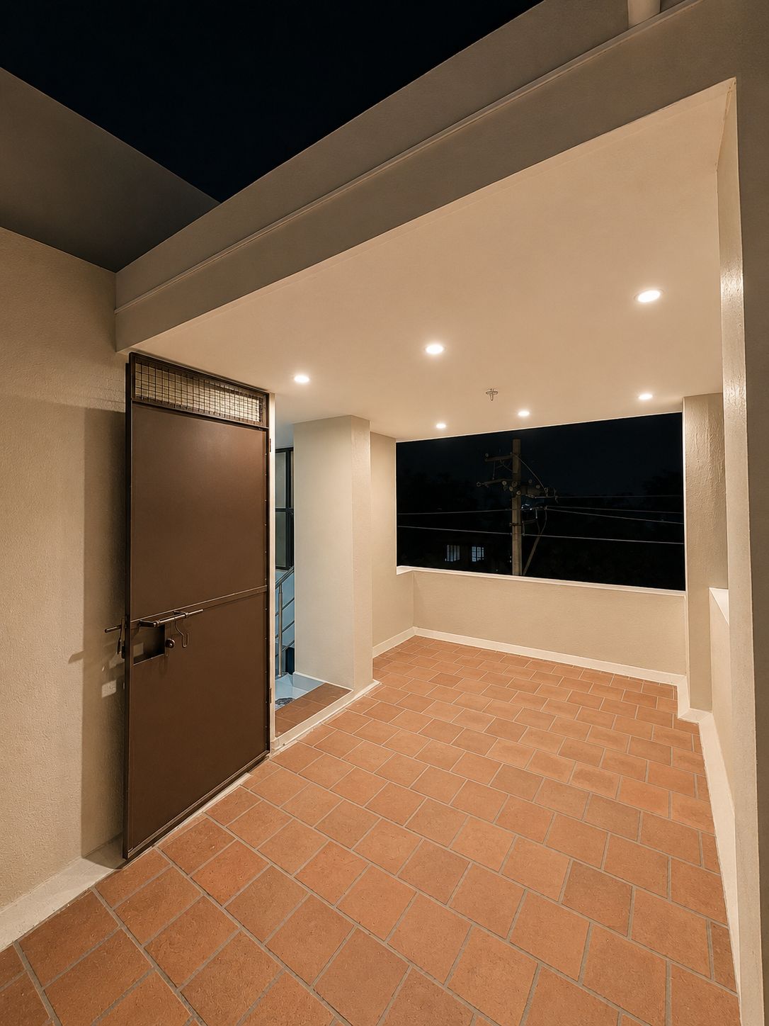 Un balcón con piso de baldosas, una puerta cerrada con reja metálica y vista al cielo nocturno.