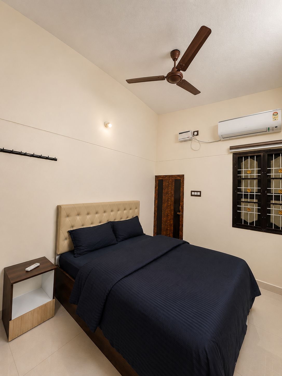 Un dormitorio con cama, mesita de noche, ventilador de techo, unidad de aire acondicionado y ventana con celosías decorativas.