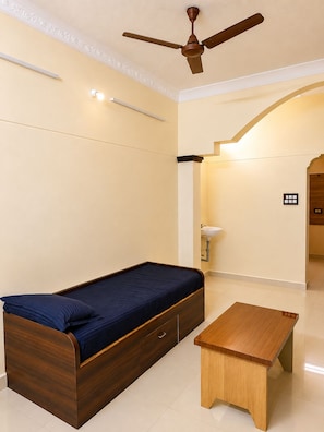 A bedroom with a single bed, a wooden nightstand, a ceiling fan, and a wall-mounted light fixture. - Sri Abirami Homestays - Residency (Tiruvannamalai)