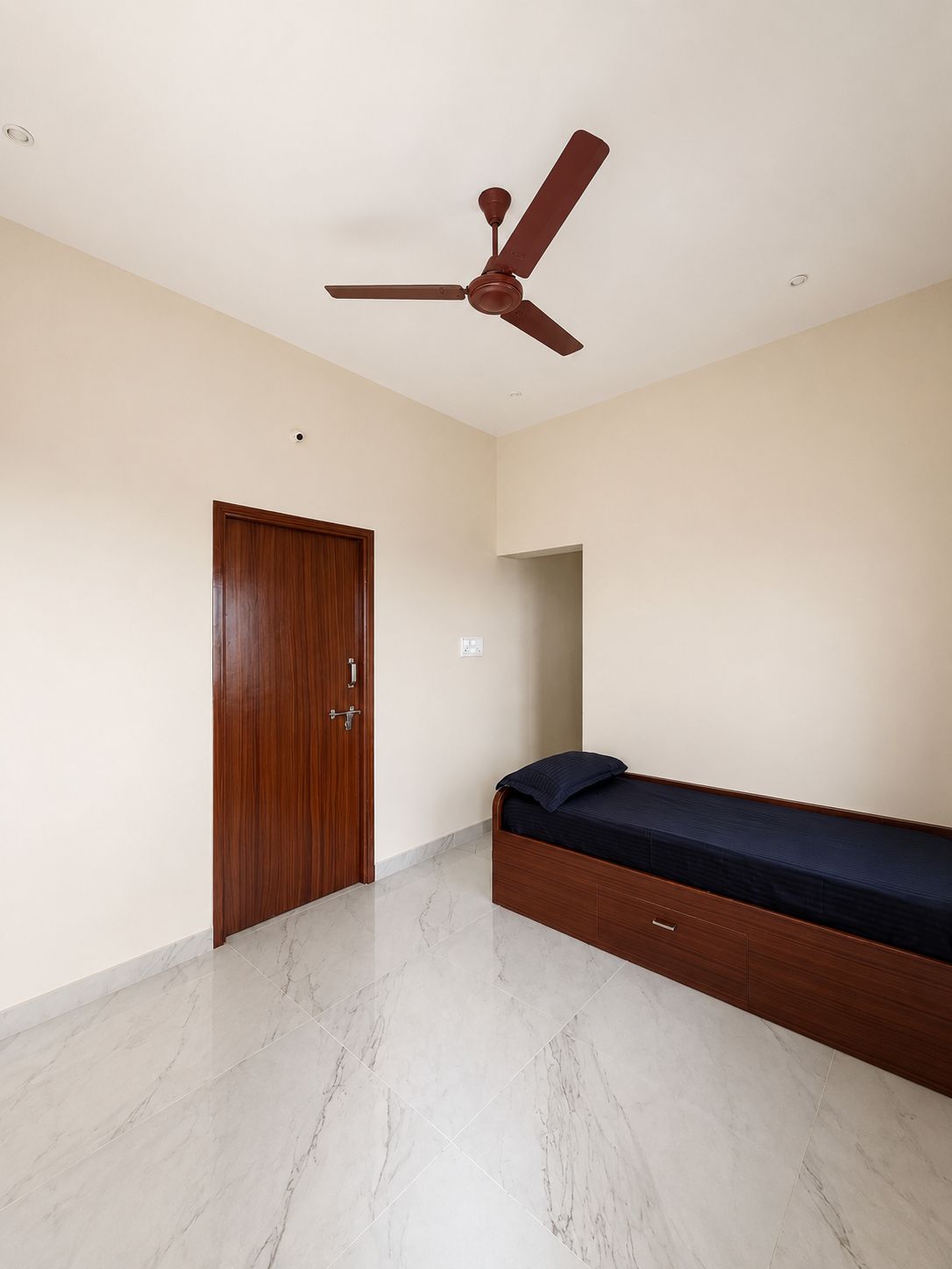 Un dormitorio con puerta de madera, una cama con colchón oscuro y un ventilador de techo.