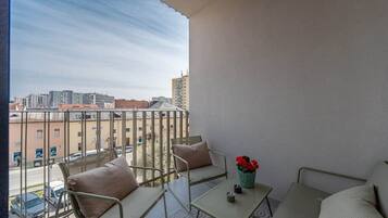 A balcony with a view of a cityscape, featuring a sofa, a table, and a vase with flowers.