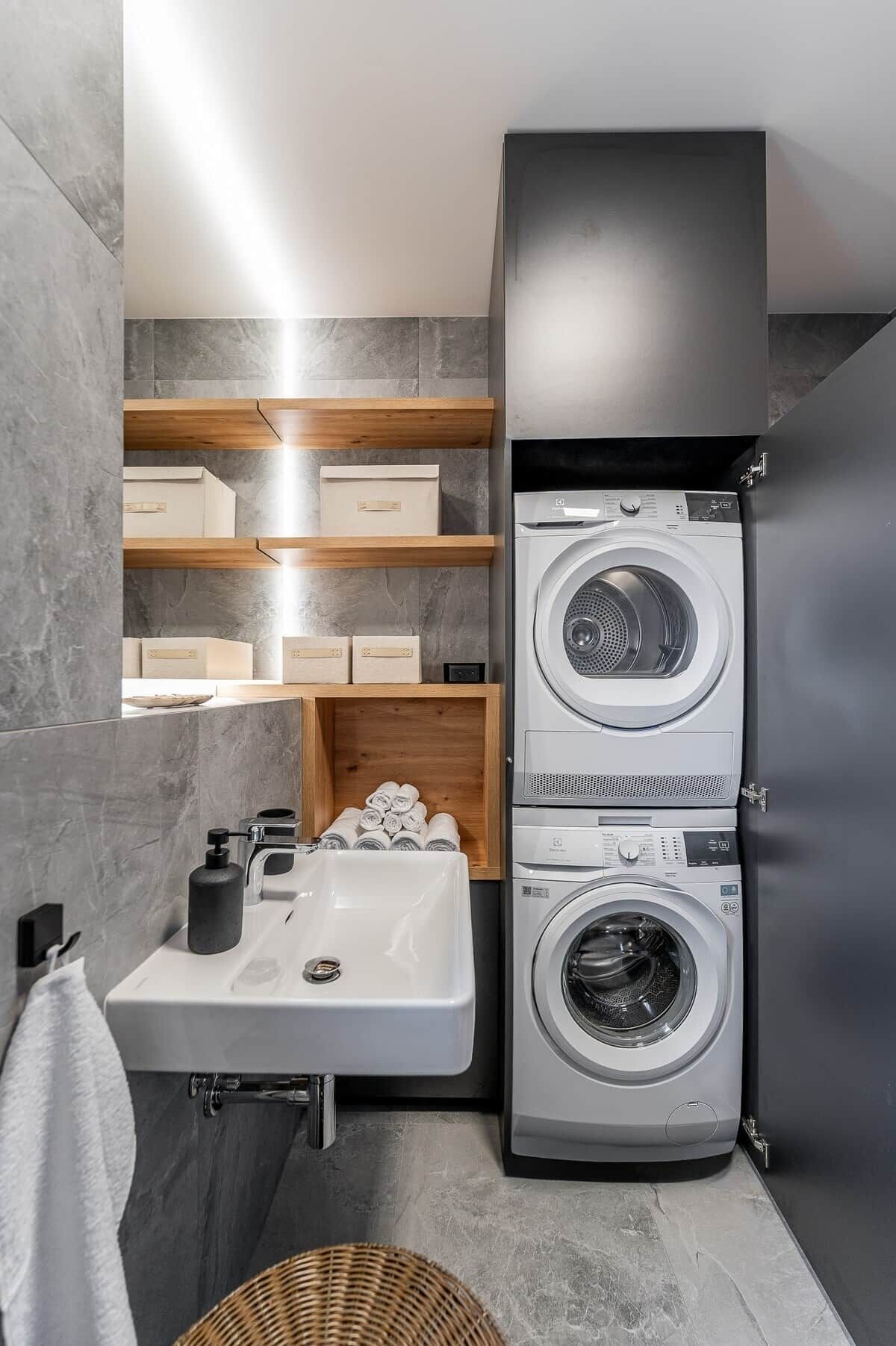 Un lavadero pequeño con lavadora y secadora frontales, un lavabo y estanterías de madera.