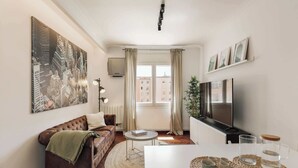 A modern living room with a sofa, a television, a dining table, and a large cityscape painting. - Enjoy Apartments Paz - Bright apartment near Sagrada Familia (Barcelona)