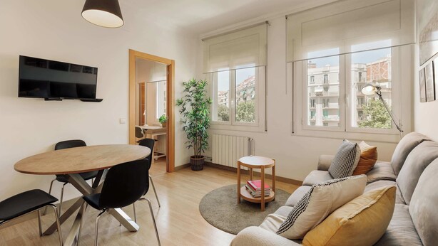 A modern living room with a flat-screen TV, a dining table with chairs, a sofa with cushions, a potted plant, and large windows with a view of buildings. - Enjoy Apartments Paz 2 - Bright apartment near Sagrada Familia (Barcelona)