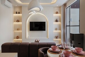 A modern living room with a sofa, a flat-screen TV mounted on the wall, and a dining area with pink plates and glasses. - Kamala Villa by Betterplace (Pecatu)
