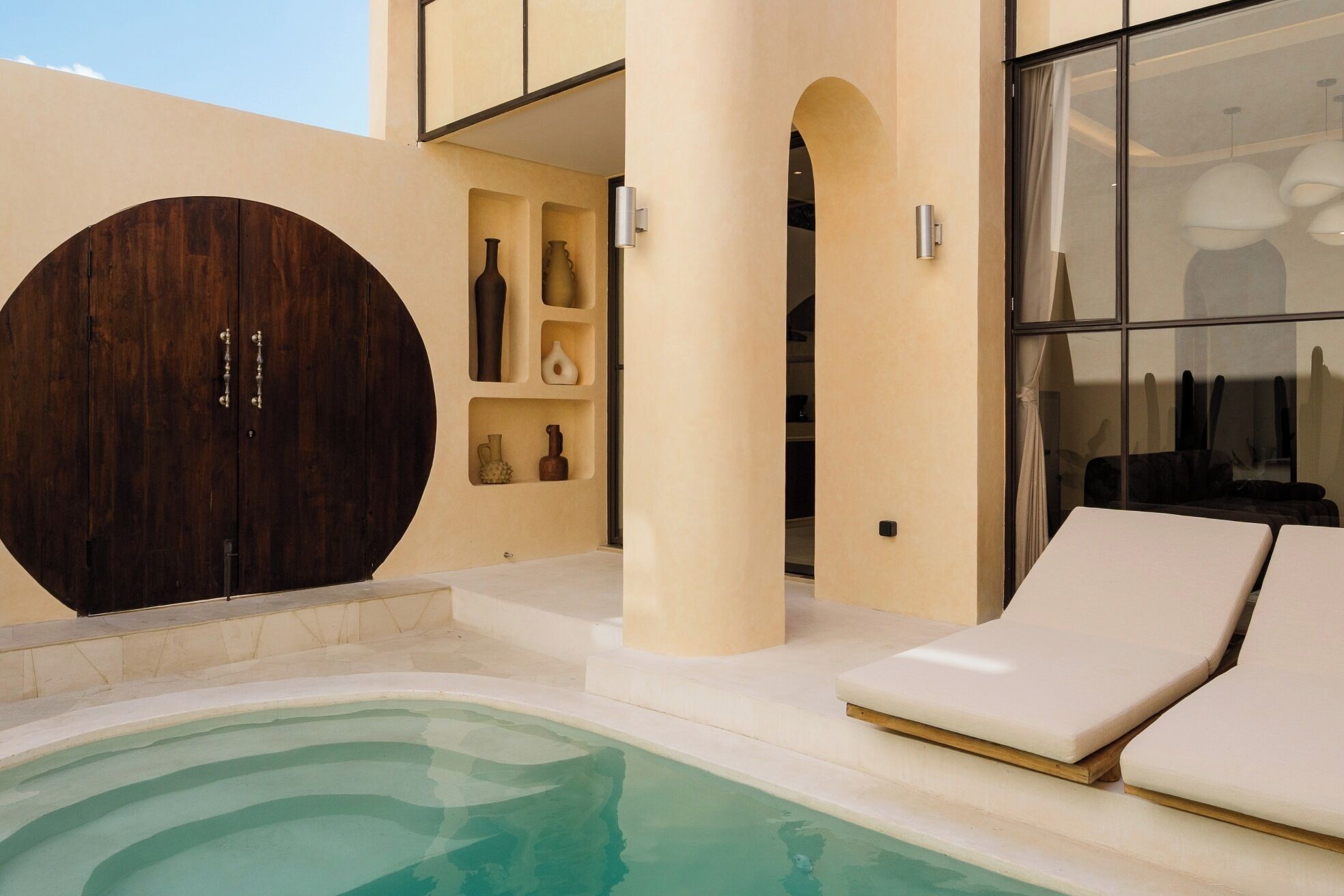 A modern outdoor pool area with a lounge chair, a large wooden door, and a glass wall.