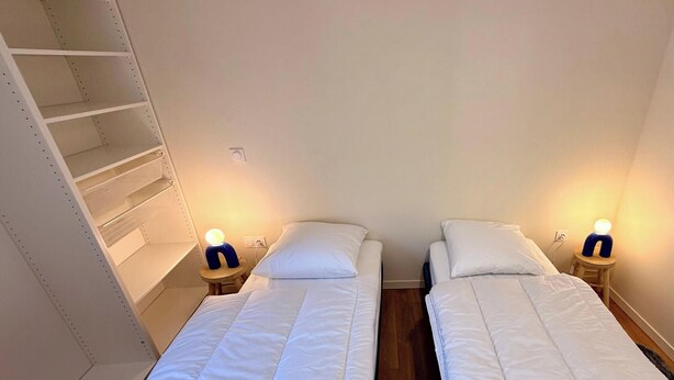 A room with two single beds, each with a bedside table and a lamp. There is a shelving unit on the left wall. - Le Belvédère de La Montille (sleeps 6) (Mont-Dore)