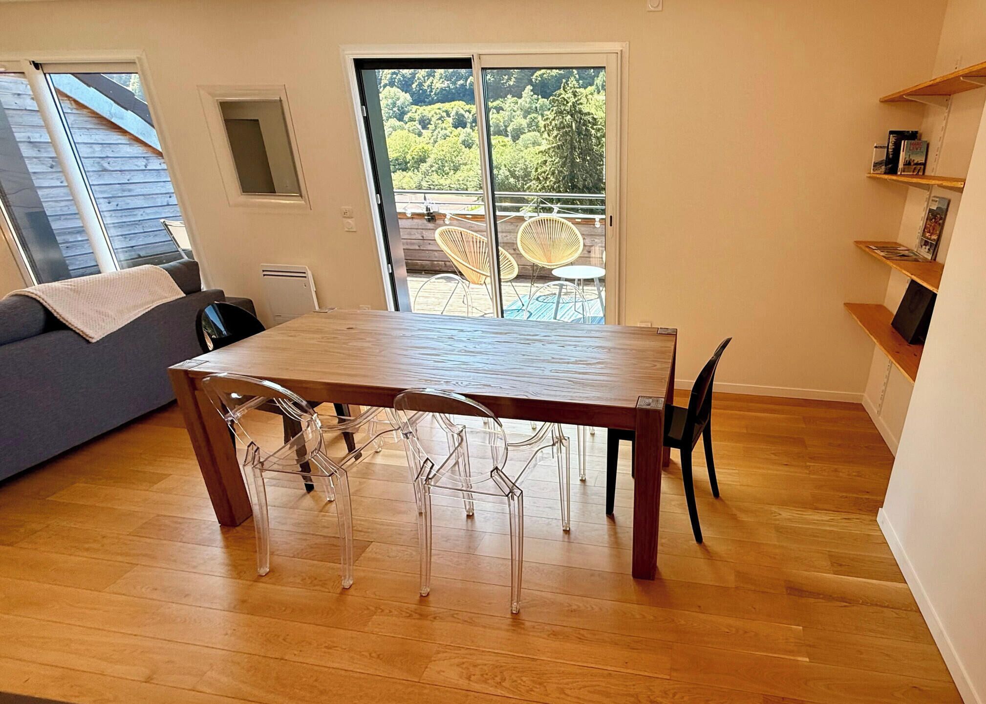 Et spiseområde med et træbord og klare stole, en grå sofa og en skydedør af glas, der fører til en udendørs terrasse.