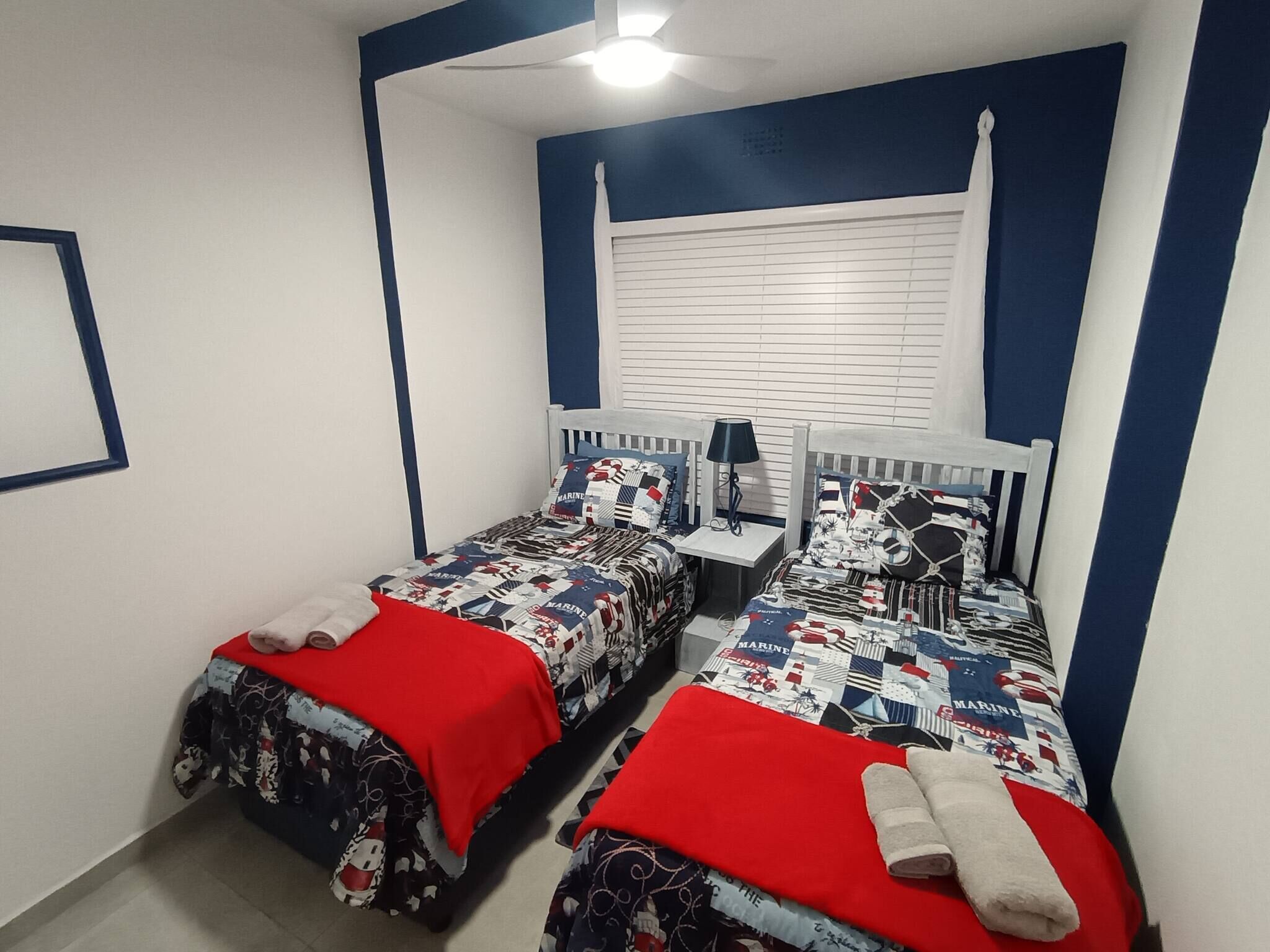 A bedroom with two beds, each with a red blanket and a marine-themed duvet. There is a nightstand with a lamp between the beds, and a window with blinds.