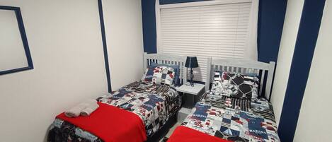 A bedroom with two beds, each with a red blanket and a marine-themed duvet. There is a nightstand with a lamp between the beds, and a window with blinds.