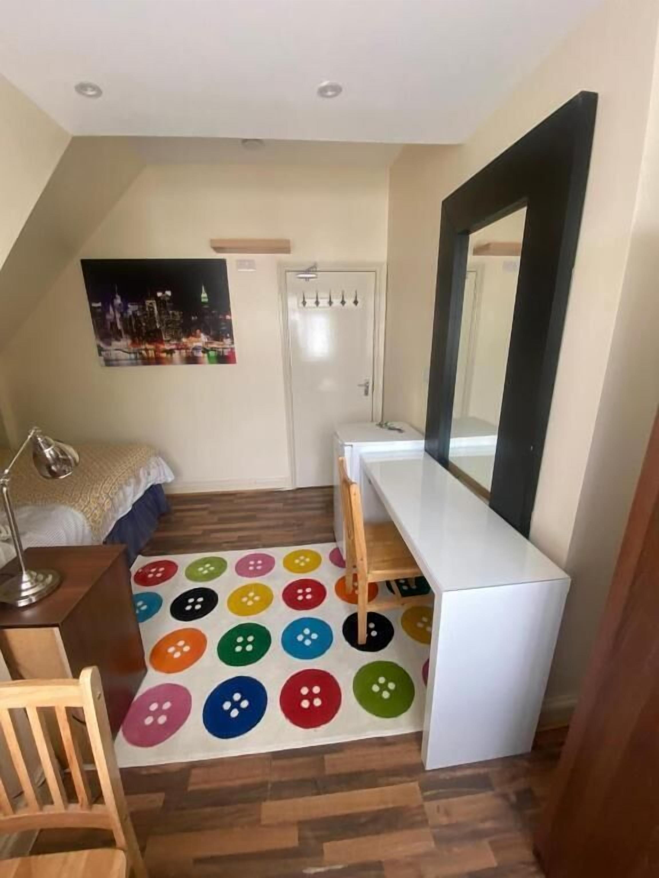 A bedroom with a bed, a desk, a mirror, a colorful rug, and a door.