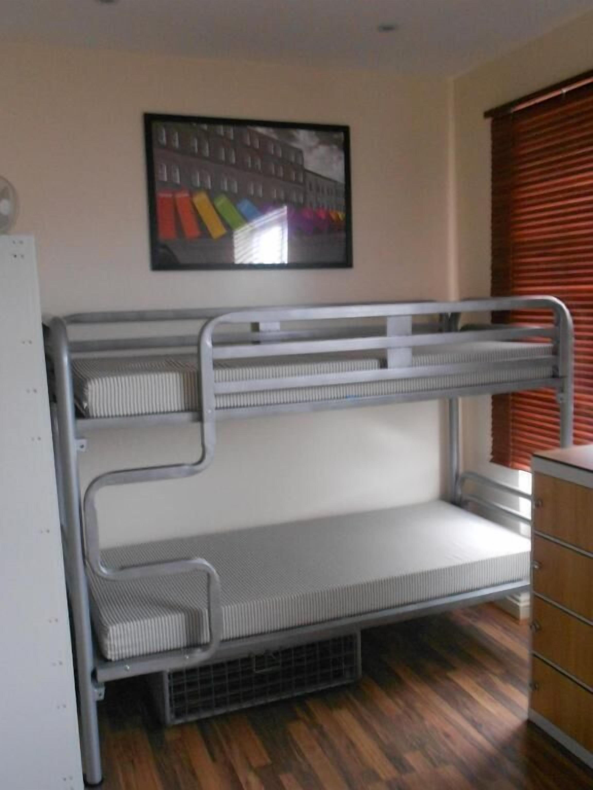 Een stapelbed met metalen spijlen, een klein houten dressoir en een ingelijste foto aan de muur.