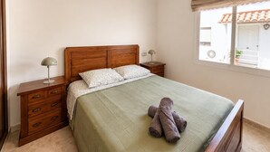 A bedroom with a wooden headboard, a green bedspread, two bedside lamps, a wooden nightstand with drawers, and a window with a view of a building outside. - The tranquility of the Mediterranean, the comfort of a spacious apartment in Villajoyosa (La Vila Joiosa)