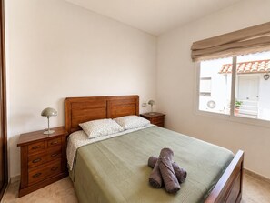 Ein Schlafzimmer mit hölzernem Kopfteil, einer grünen Bettdecke, zwei Nachttischlampen, einem Nachttisch mit Schubladen und einem Fenster mit Blick auf ein Gebäude.