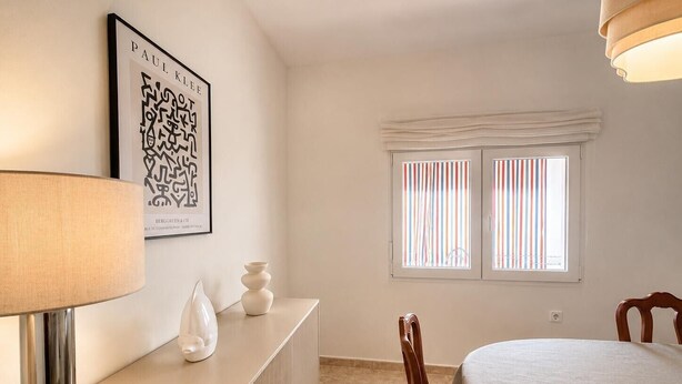 A room with a bed, a bedside table with a lamp, a decorative vase, a framed artwork, and a window with striped blinds. - The tranquility of the Mediterranean, the comfort of a spacious apartment in Villajoyosa (La Vila Joiosa)