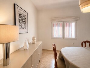 Una habitación con cama, una mesita de noche con lámpara, un jarrón decorativo, una obra de arte enmarcada y una ventana con persianas a rayas.