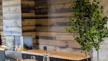 Een eettafel gedekt met kaarsen, borden en glazen, met een houten wand en een plant op de achtergrond.