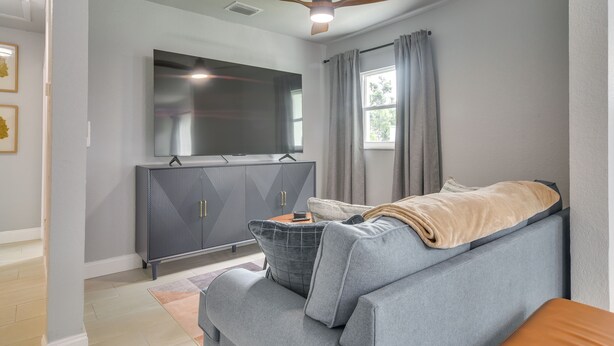 Ein modernes Wohnzimmer mit einem grauen Sofa, einem flachbildfernseher an einer Kommode, einem Deckenventilator und einem Fenster mit Vorhängen.