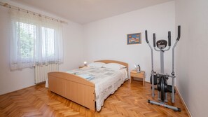Ein Schlafzimmer mit einem hölzernen Bett, einem Nachttisch, einer Lampe, einem Fenster mit Vorhängen und einem stationären Heimtrainer.