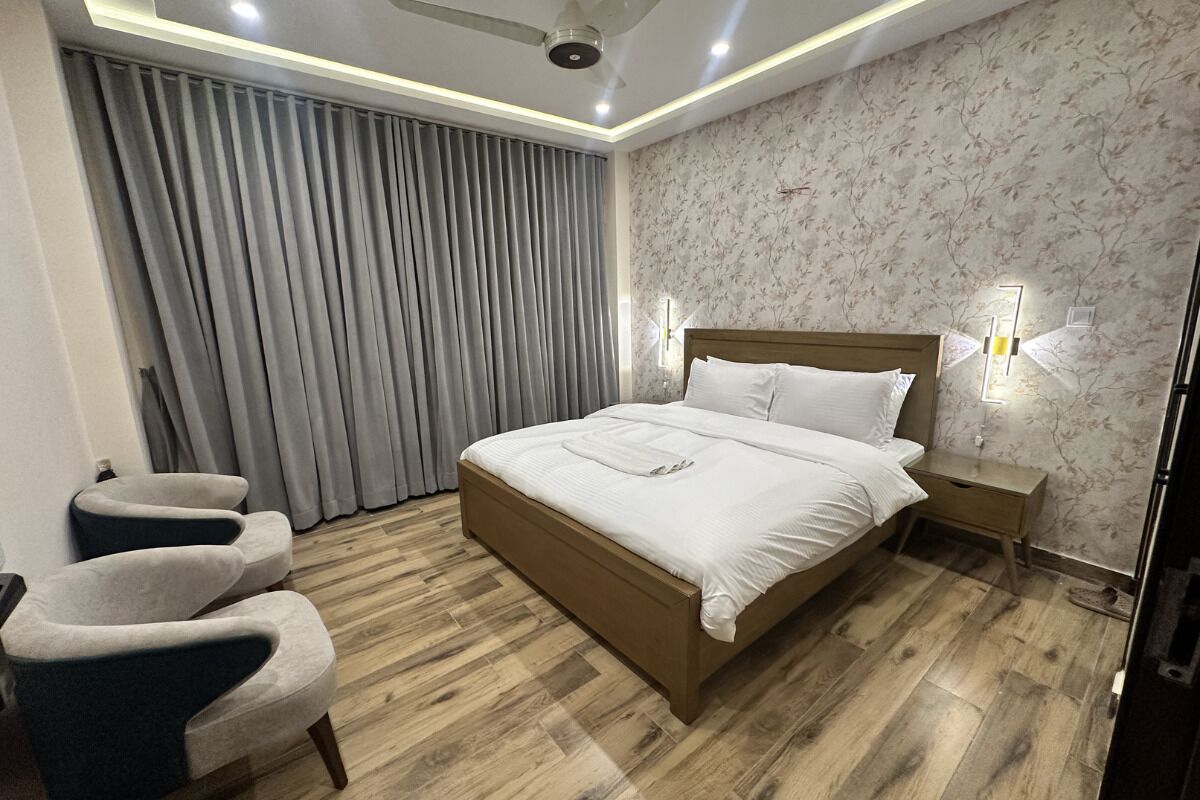 Quarto moderno com cama de madeira, duas poltronas, mesa de cabeceira e uma janela grande com cortinas.