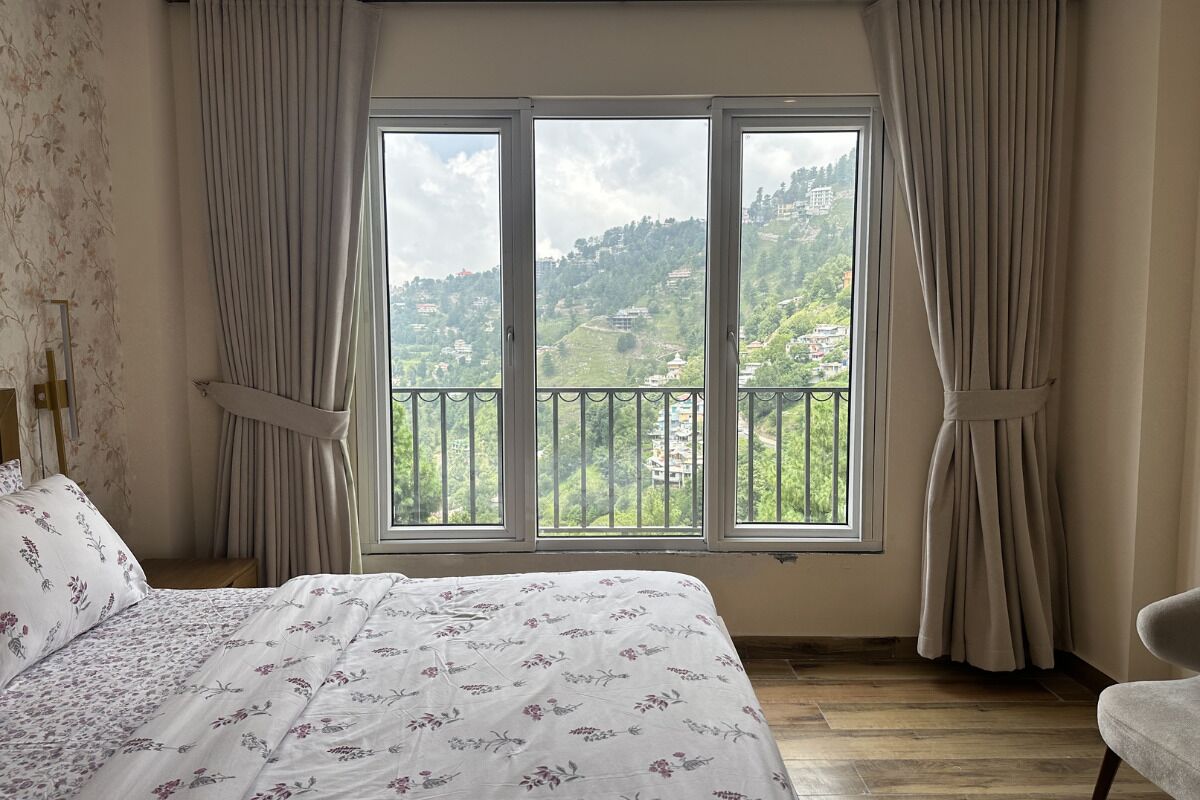 Une chambre avec une grande fenêtre, un lit recouvert d’une literie à motifs floraux et une vue sur un paysage verdoyant.