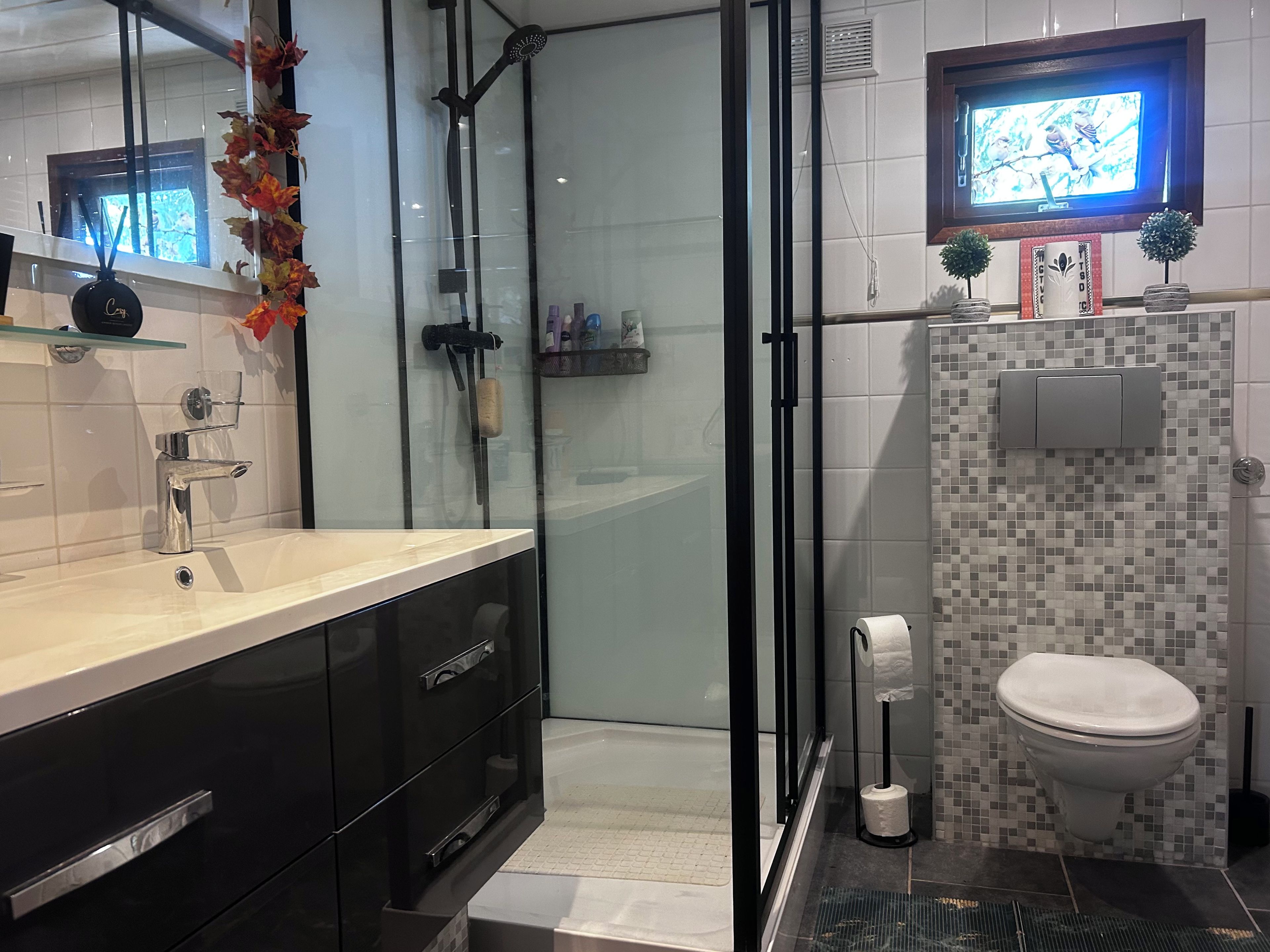 Une salle de bain moderne dotée d’une douche à parois de verre, de toilettes décorées de mosaïques et d’un meuble-lavabo avec des armoires noires.