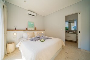 A bedroom with a bed, bedside lamps, a wall-mounted air conditioner, and a bathroom visible through an open door. - Calula Beach House II Uluwatu (Uluwatu)