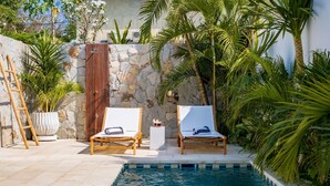 Área junto à piscina com duas espreguiçadeiras, um muro de pedra e plantas tropicais.