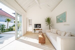A modern living room with a white sofa, a wooden coffee table, a TV, and a large window. - Calula Beach House II Uluwatu (Uluwatu)