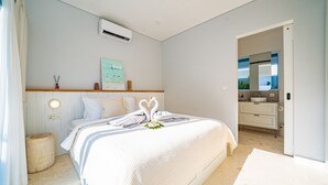 Quarto com cama, mesa de cabeceira, ar condicionado, prateleira na parede com objetos decorativos e banheiro visível por uma porta aberta.