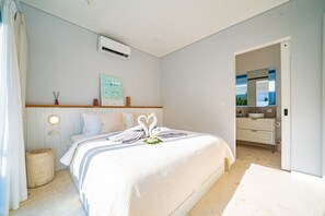 Ein Schlafzimmer mit Bett, Nachttisch, Klimaanlage, einer an der Wand befestigten Ablage mit Dekoration und einem durch eine offene Tür einsehbaren Badezimmer.