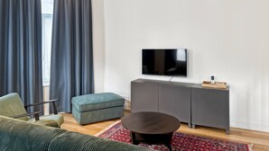 En moderne stue med en sofa, sk foot og et rundt sofabord. Et fladskærms-tv er monteret på væggen, og der er et gråt skab nedenunder.