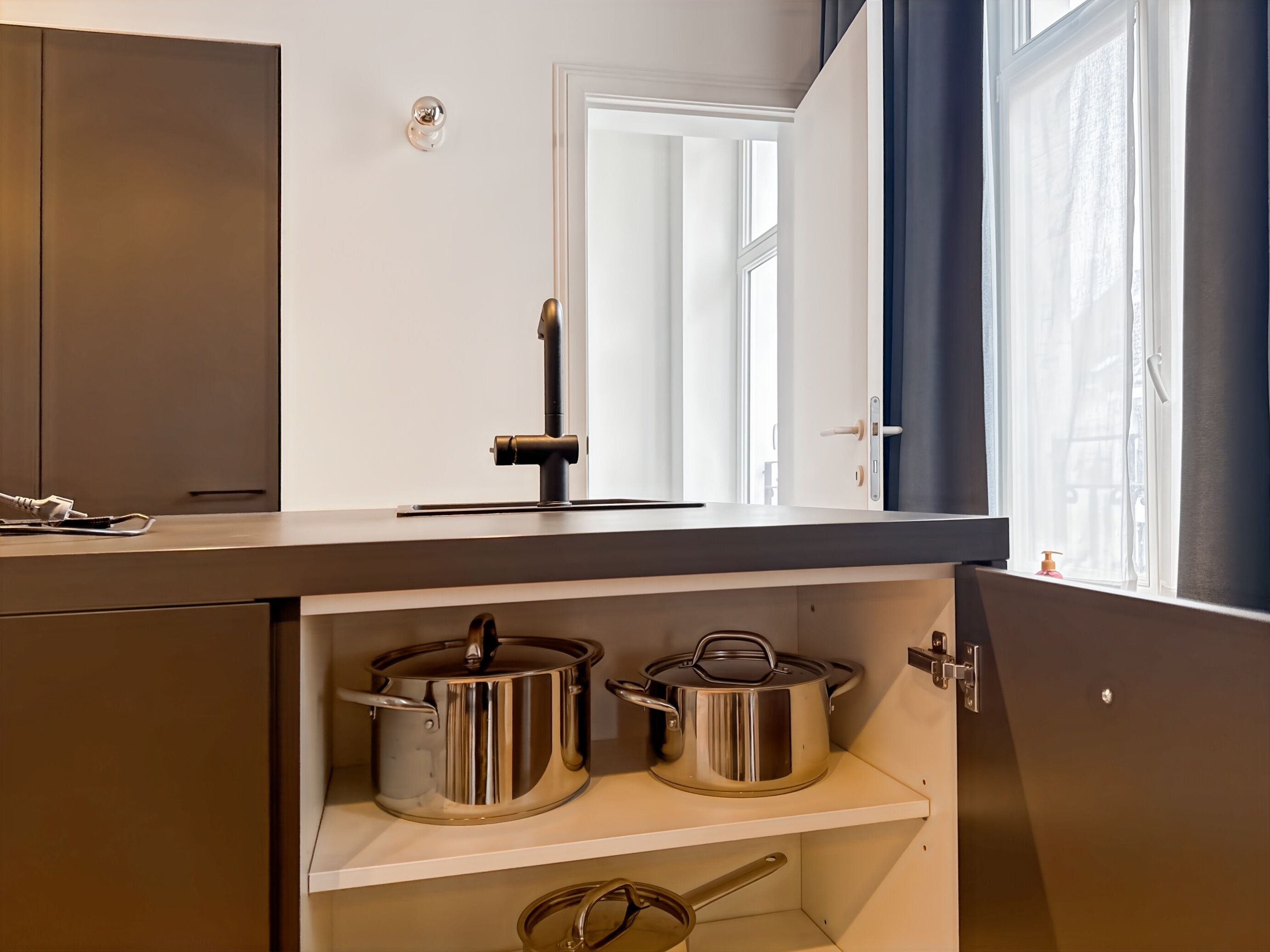 A kitchen with a built-in stove, two stainless steel pots, and a window with curtains.