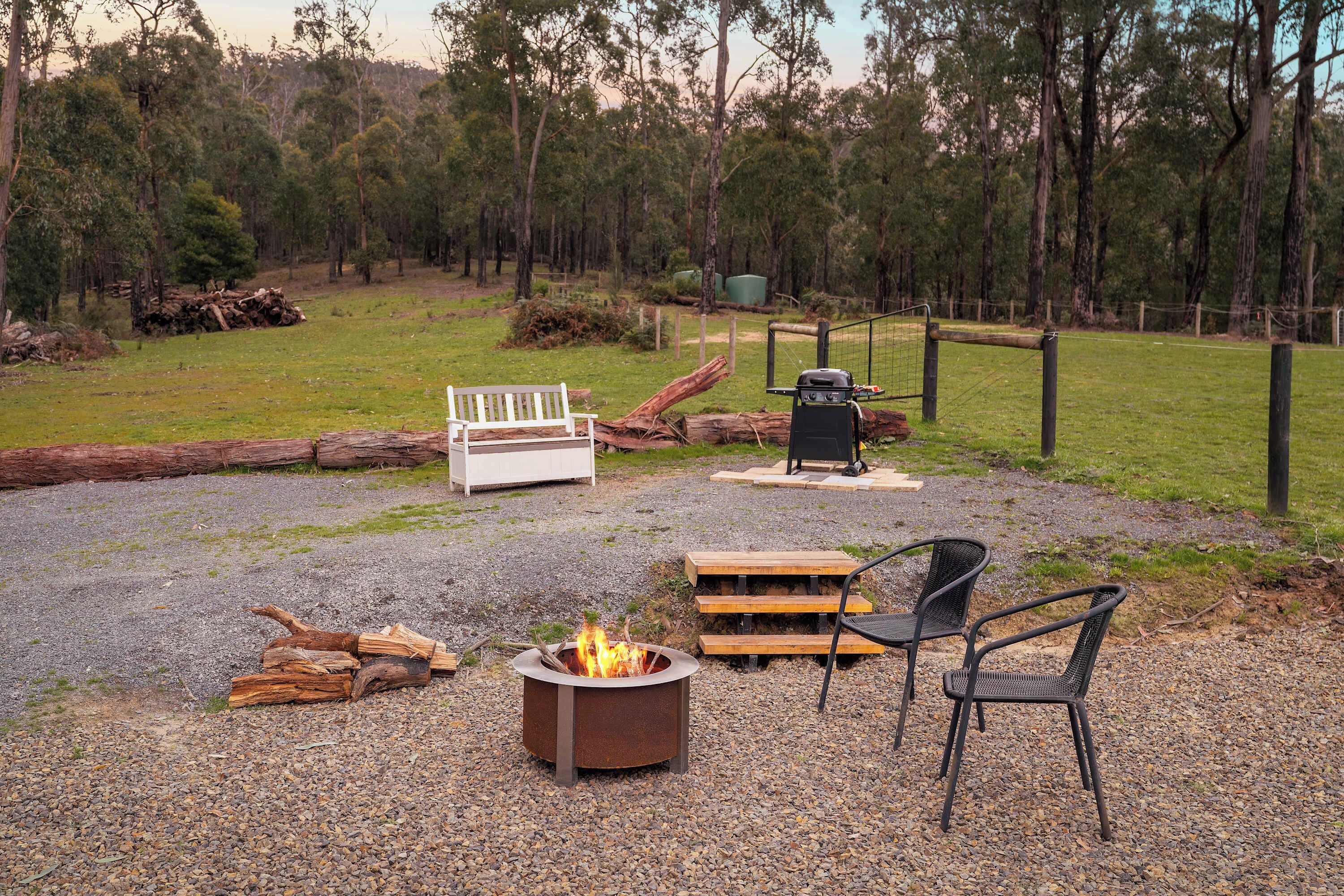 A gravel patio with a fire pit, a bench, a grill, and a chair.