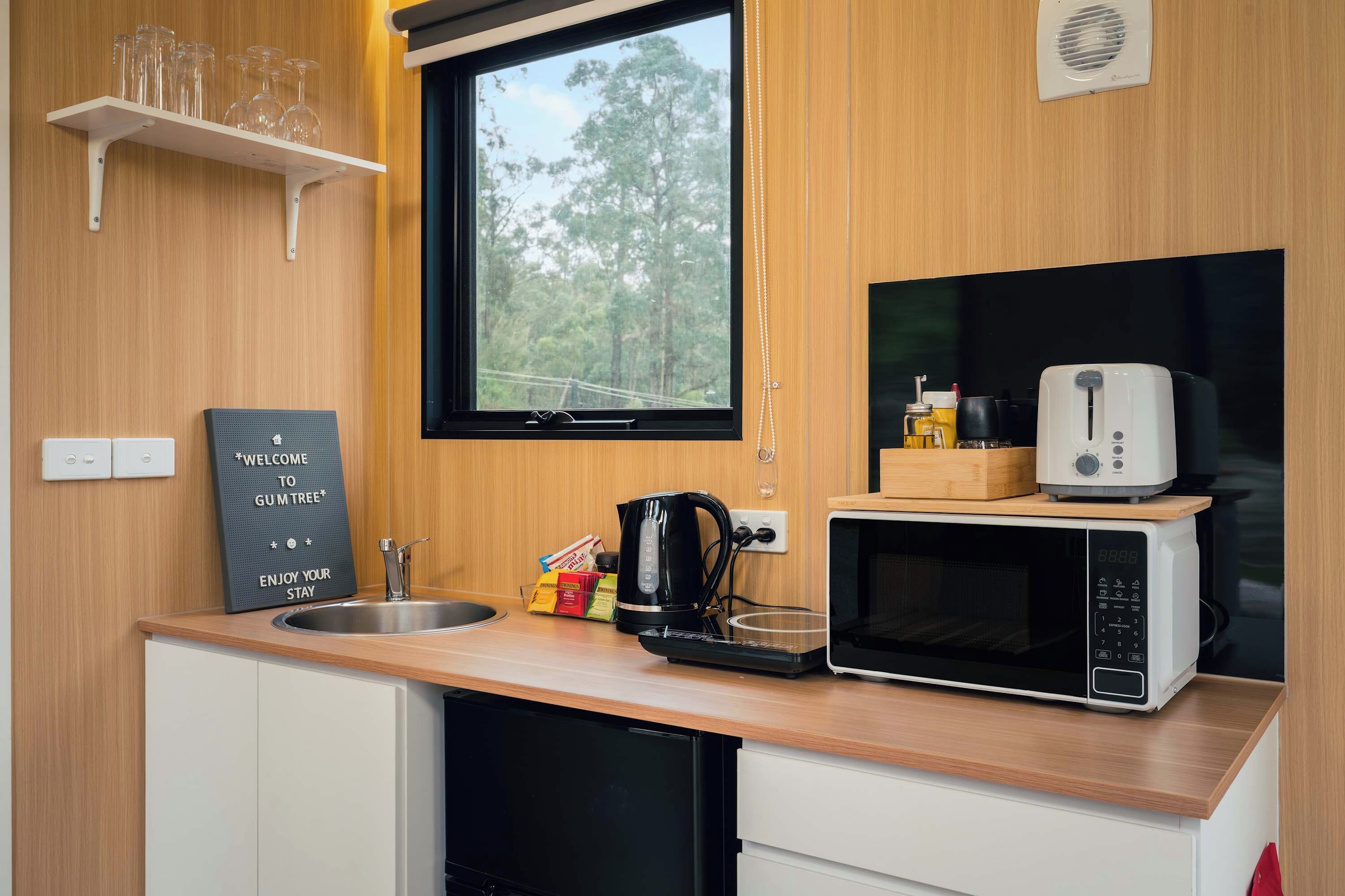 A compact kitchenette with a microwave, kettle, and a small coffee maker. There is a window above the sink, and a welcome sign is visible on the wall.