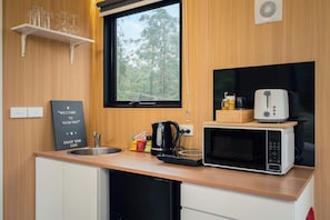 A compact kitchenette with a microwave, kettle, and a small coffee maker. There is a window above the sink, and a welcome sign is visible on the wall.