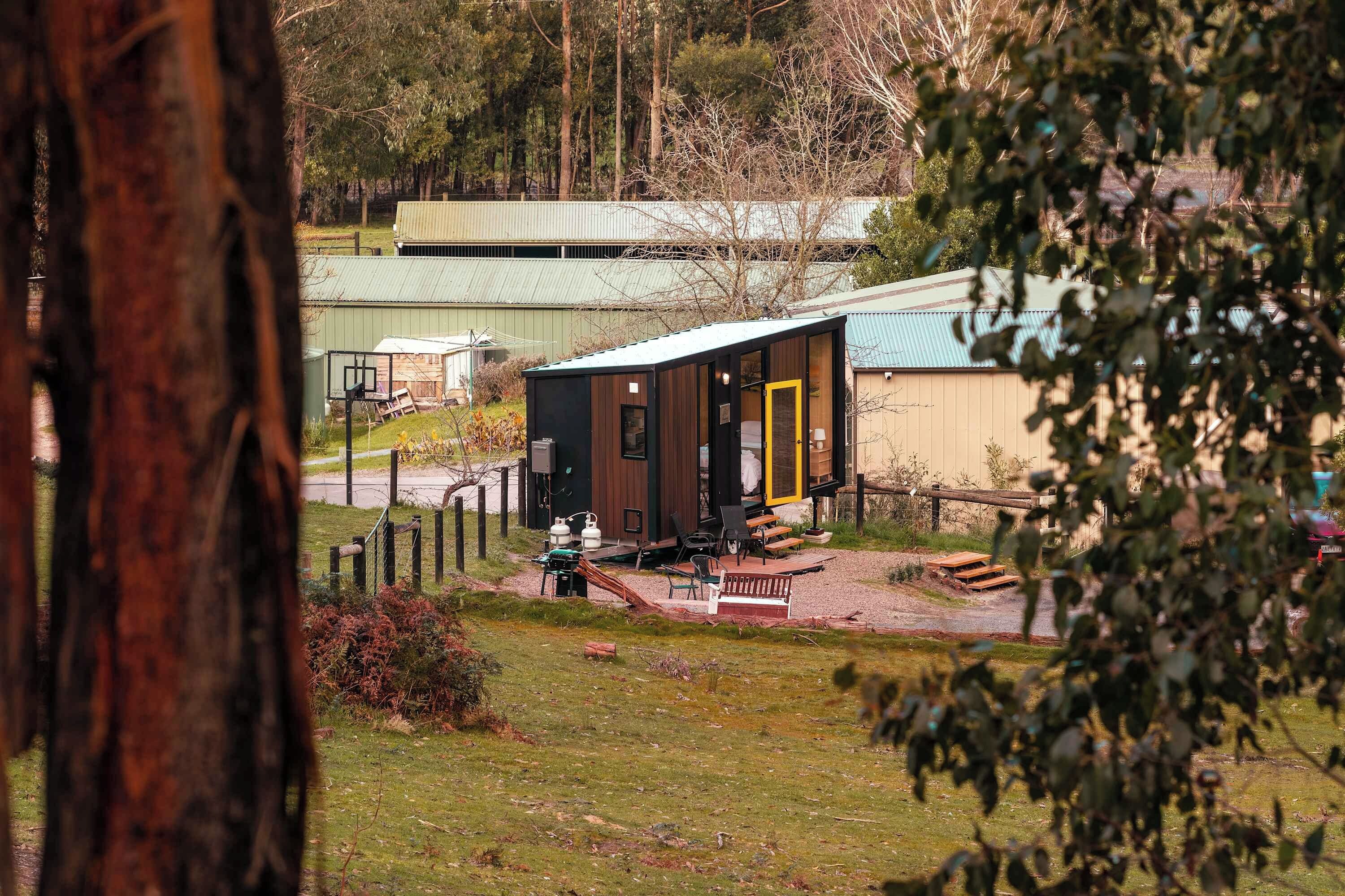 A small cabin with a deck, surrounded by a fence, and a larger building in the background.