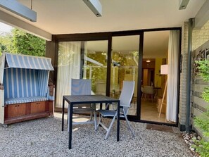 Un espace extérieur aménagé avec une table et des chaises, un auvent à rayures et une vue sur l’intérieur de la maison à travers une porte vitrée.