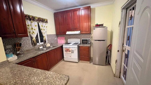 A compact kitchen with granite countertops, wooden cabinets, a stove, microwave, and refrigerator. - A beautiful, comfortable, and safe home for the whole family (Santo Domingo Este)