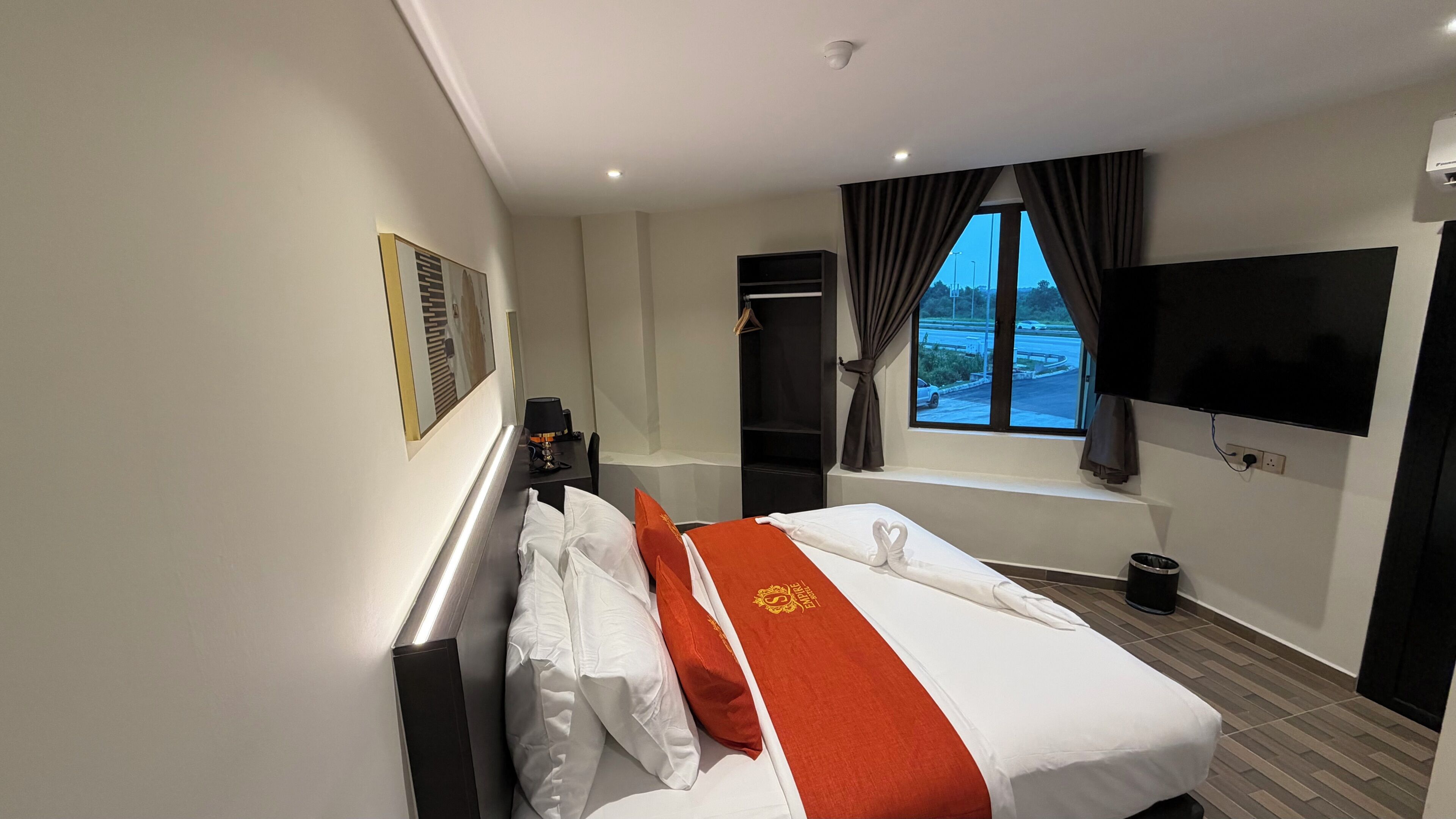 A hotel room with a bed, a TV, a window with curtains, a coat rack, and a wall-mounted light fixture.