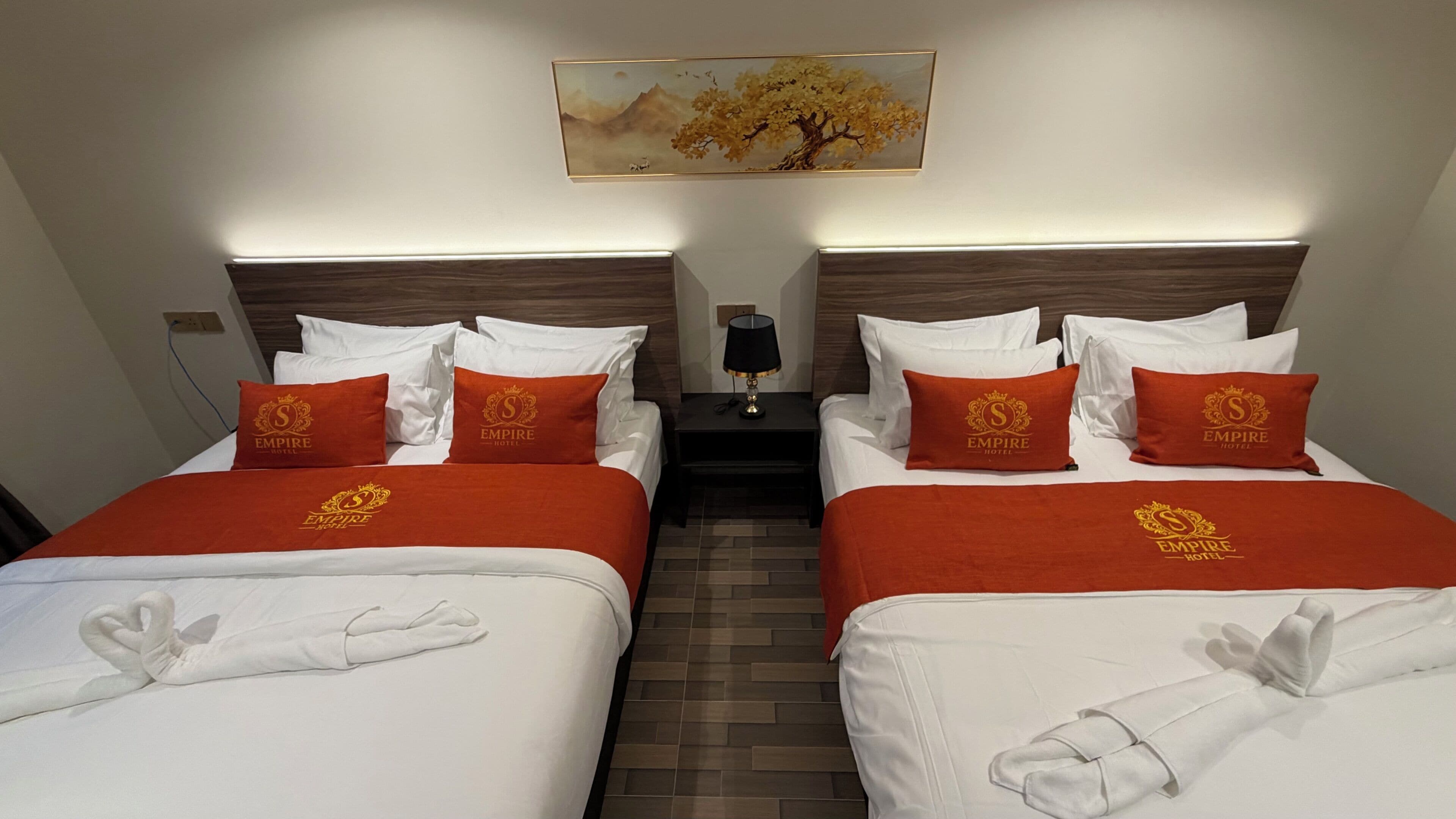 A hotel room with two beds, each with a red Empire Hotel logo pillow, white bedding, and a decorative swan towel. A nightstand with a lamp and a painting of a tree and mountains is centered above the beds.